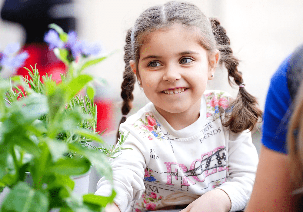 A child with a joyful expression looks at plants, wearing a floral and text-patterned top.