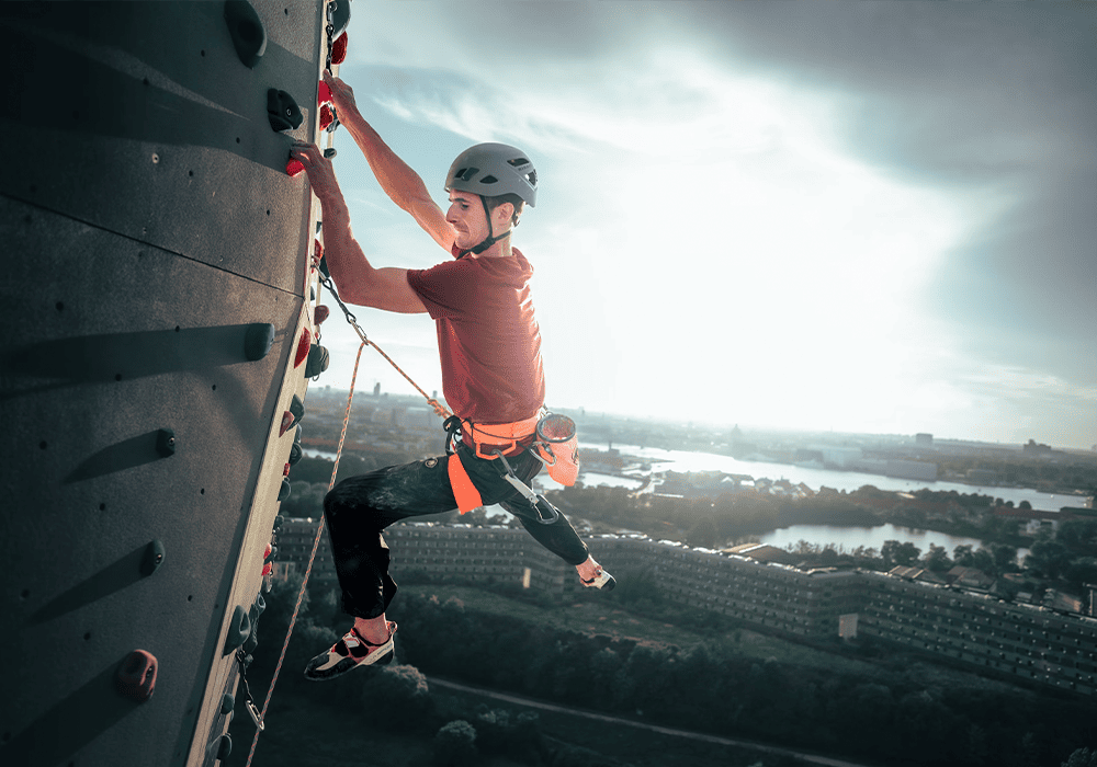 Adam Ondra climbing an outdoor artificial rock wall with a scenic view of a river and cityscape in the background, wearing a helmet and safety harness.