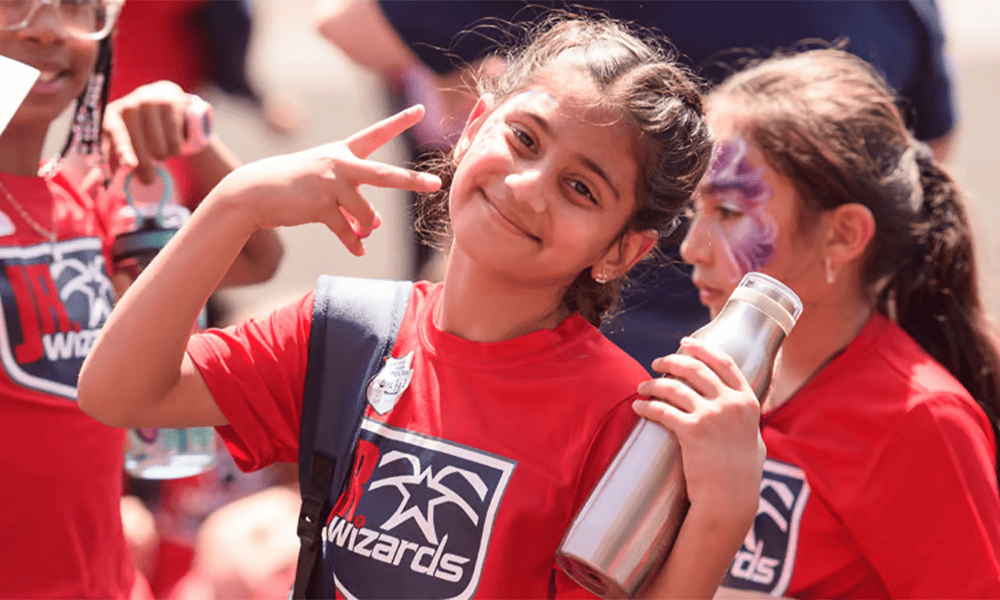 A group of children wearing Washington Wizards t-shirts, one child in the foreground smiling and gesturing a peace sign, during a sunny outdoor event.