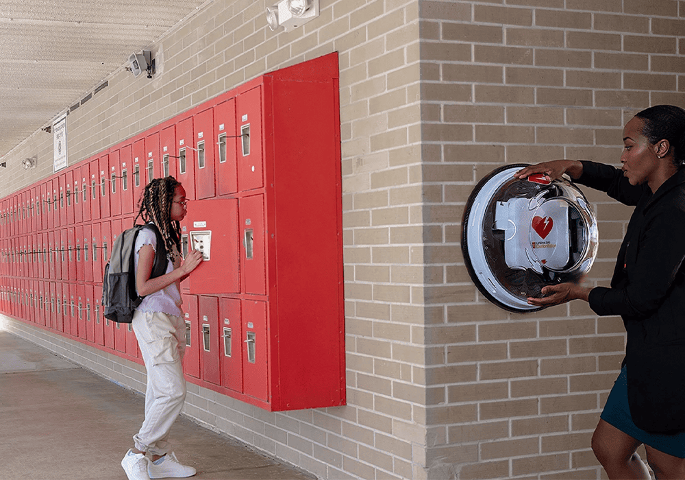 Young girl student stands next to her lockers, on the other wall a woman is attaching an AED to the wall, which is a life-saving tool against sudden cardiac arrest 