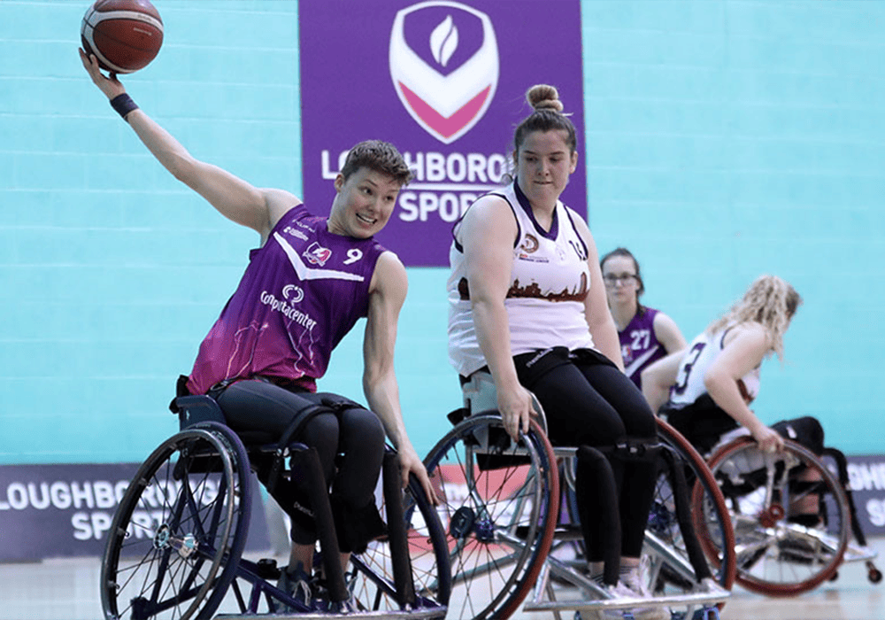 Two wheelchair basketball players in action during a game, one wearing a purple Loughborough University jersey stretching to pass the ball, while the other, in a white jersey, plays defense. They are on a court with a Loughborough Sports logo visible in the background.