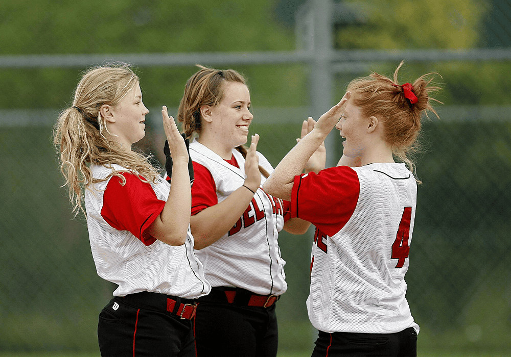 Three baseball players in white and red uniforms labeled are celebrating with high fives on a grassy field.