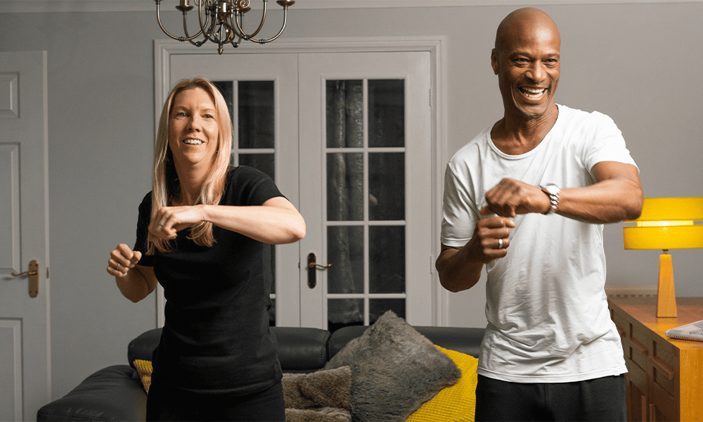 Two adults smiling and exercising in a home setting.