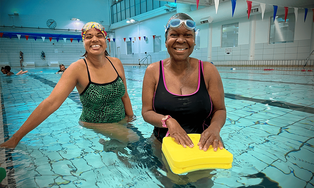 Two people smiling in a swimming pool, each holding a kickboard. One person is wearing a green swimsuit and the other a purple swimsuit. Flags hang above the water in the background.